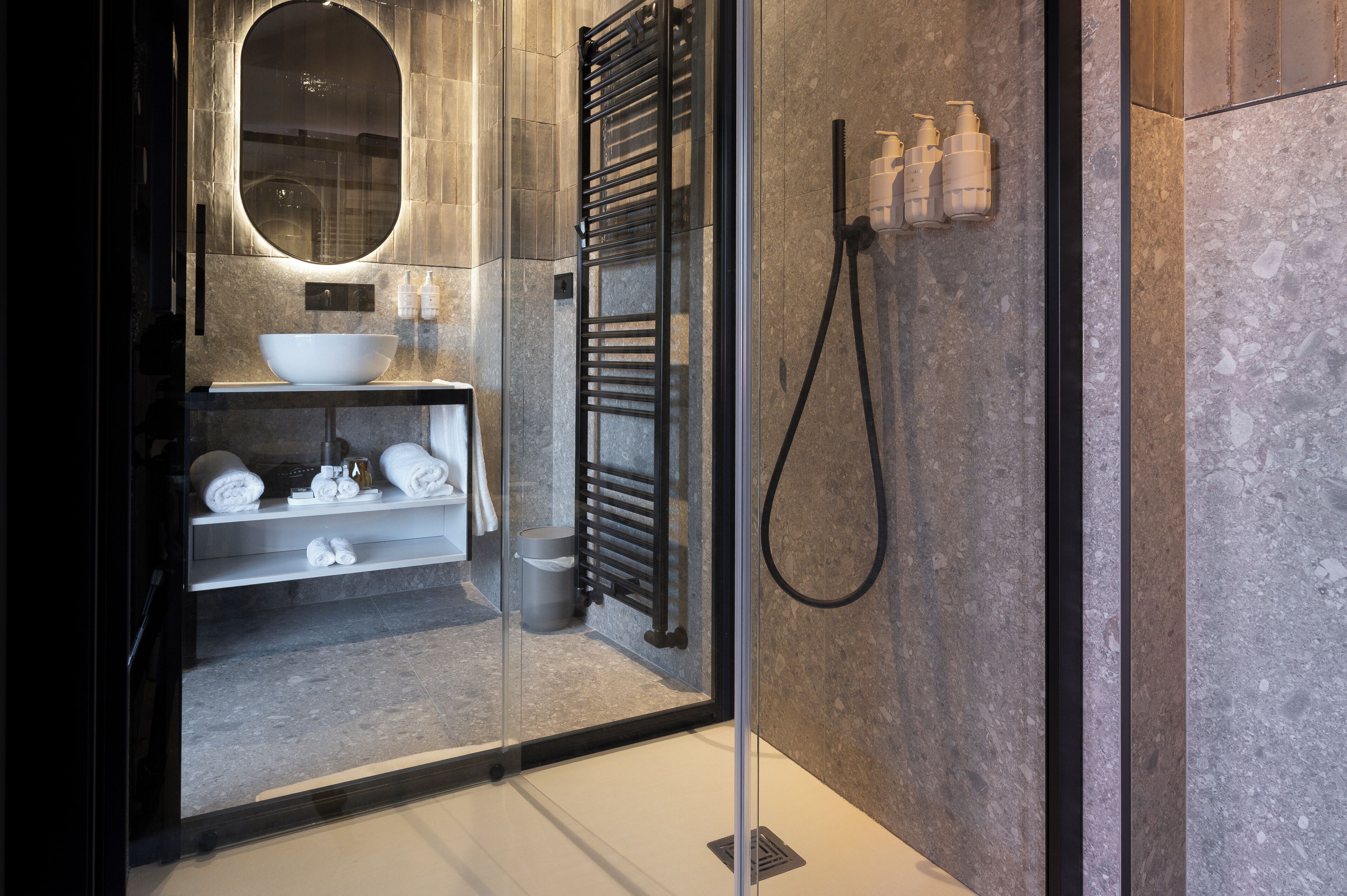 Un baño moderno con ducha envuelta en vidrio, un espejo ovalado grande, un lavabo blanco y un perchero para toallas.