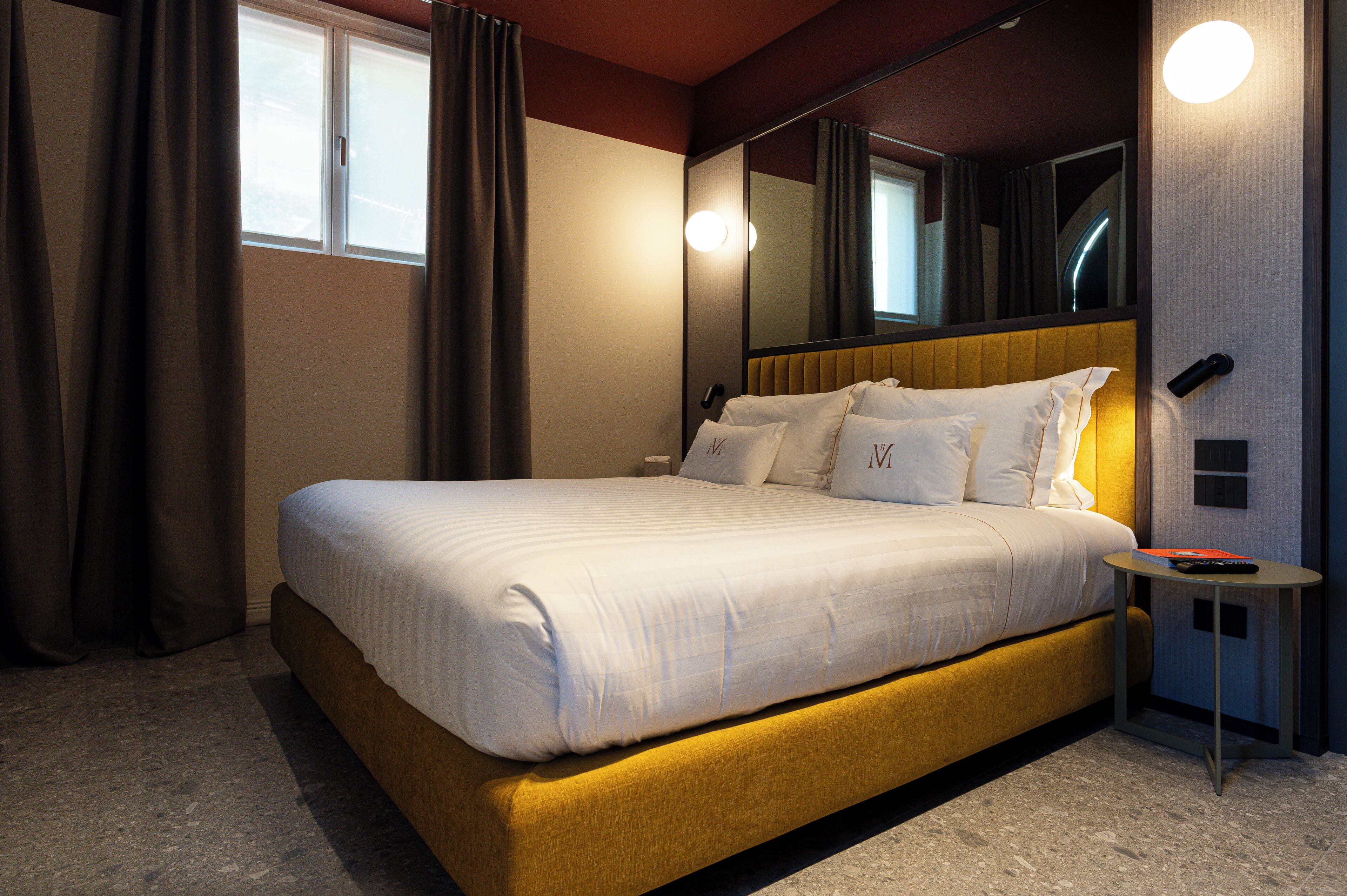 A hotel room with a large bed, a bedside table, a mirror, and a window with curtains.