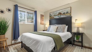 A bedroom with a large bed, a nightstand, a lamp, a painting, and a window with curtains.