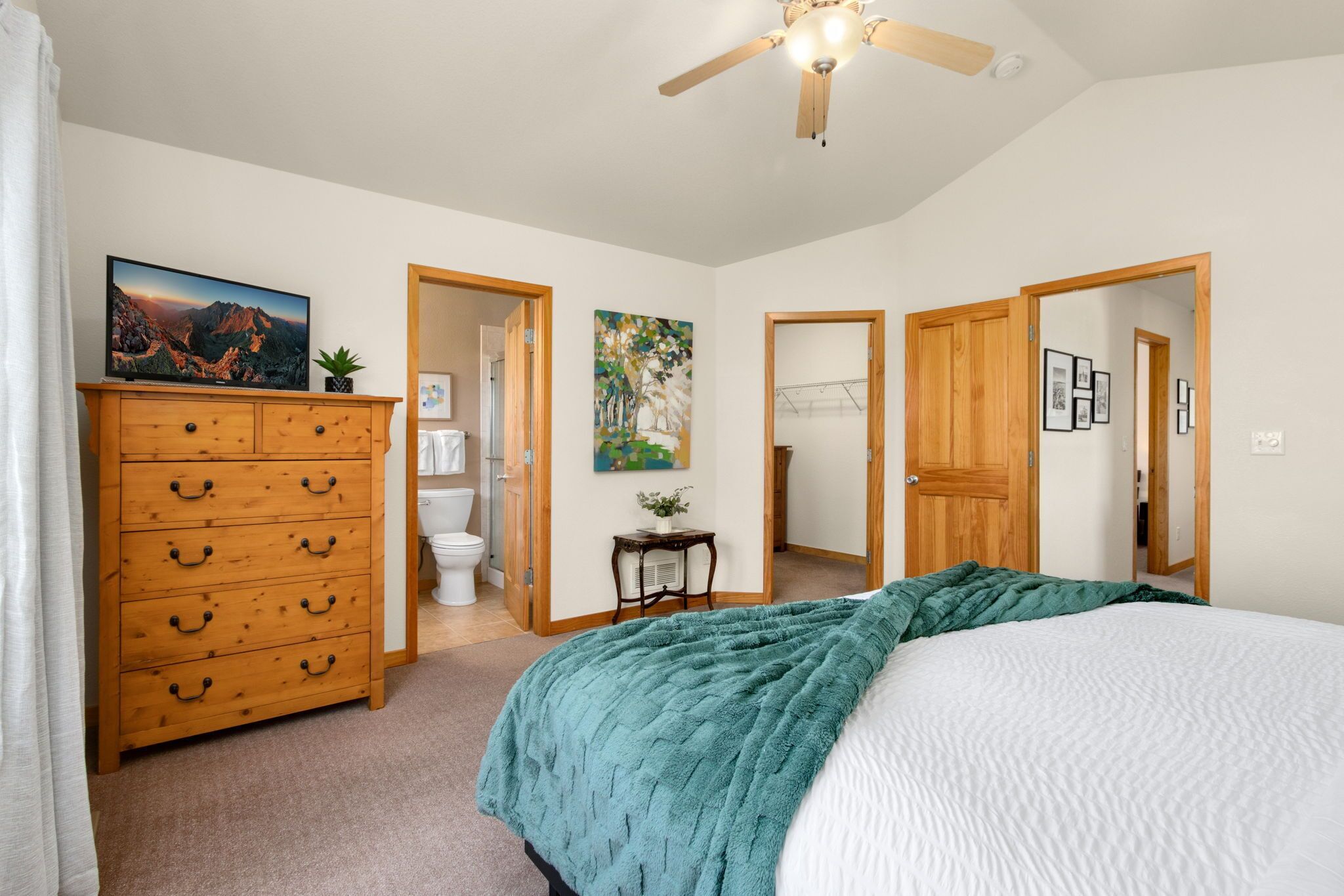 A bedroom with a bed, a wooden dresser, a ceiling fan, and a painting on the wall.