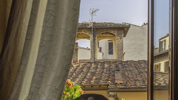 A room with a view of buildings through a window, a wicker basket with flowers, a chair, a table with a glass jar, and a cushion. - Antica Dimora De Benci (Florence)