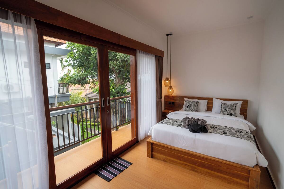 A bedroom with a wooden bed, white bedding, a nightstand, a lamp, and a view of a balcony with a railing and greenery.