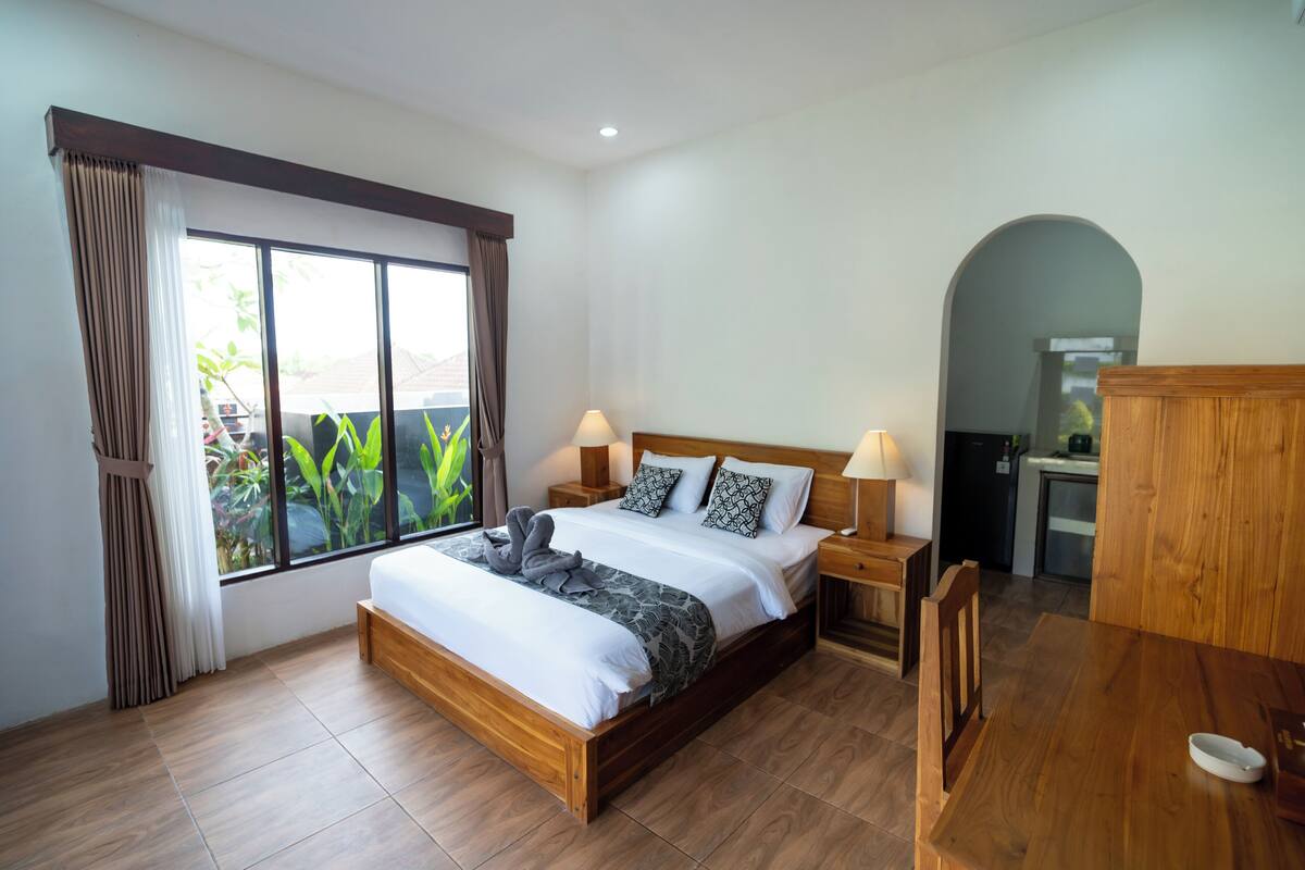A bedroom with a wooden bed, two bedside tables with lamps, a wooden desk, and a view of greenery outside.
