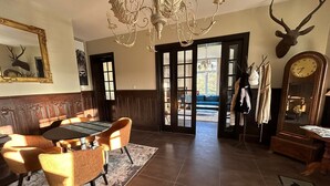 Een eetruimte met een ronde tafel en stoelen, een kroonluchter, een opgezette hertenkop aan de muur, en een grote klok.