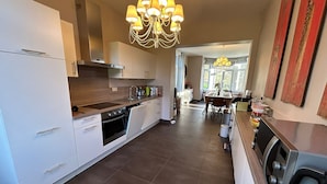 Een moderne keuken met witte kasten, roestvrijstalen apparatuur en een centraal eiland. Achtergrond toont een eethoek met tafel en stoelen.