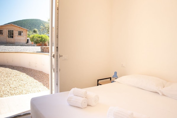 A room with a bed, a window, and a view of a building and hill.