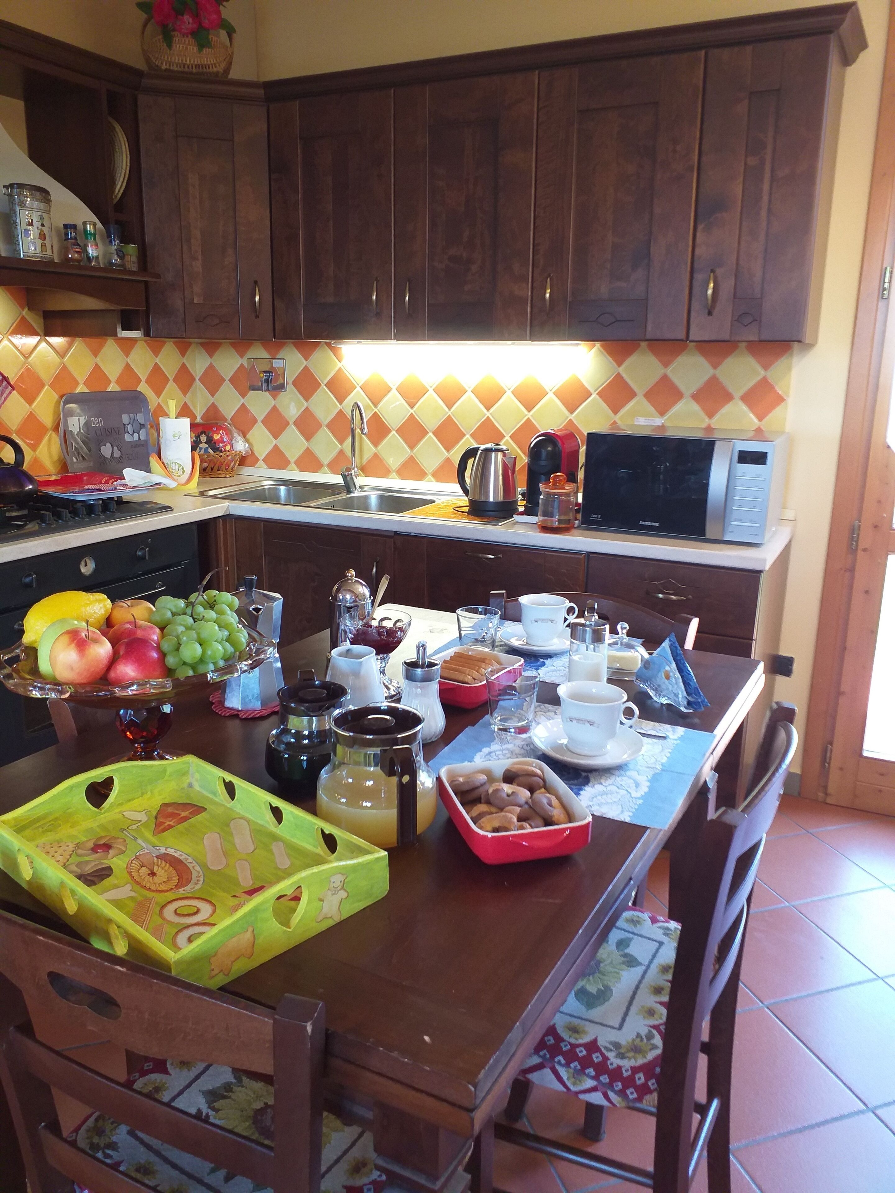 Una cocina con armarios de madera, un microondas y un lavabo. Hay un frutero, una cafetera y una mesa lista para el desayuno.