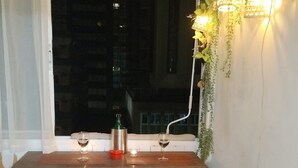 Un petit coin repas avec une table en bois et deux tabourets, une fenêtre donnant sur des immeubles et un luminaire fixé au mur, agrémenté de plantes.