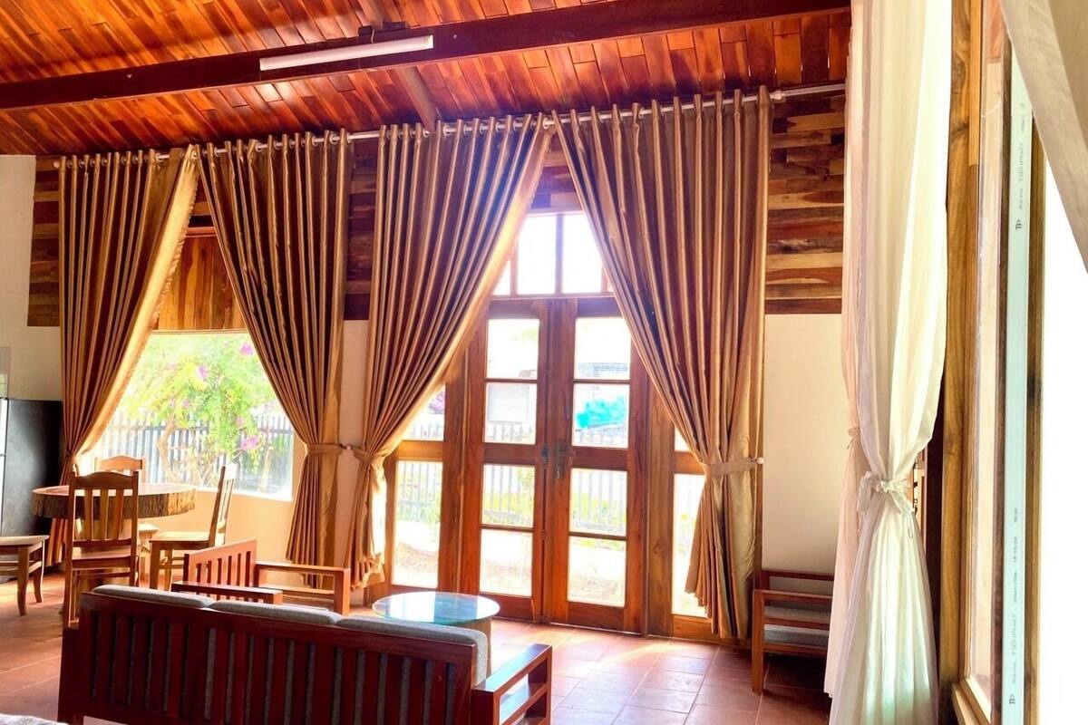 A room with a wooden ceiling, large windows with curtains, a sofa, a table, and chairs.