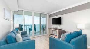 Un salon moderne avec deux canapés bleus, un téléviseur à écran plat et une porte vitrée donnant sur un balcon avec vue sur la mer.