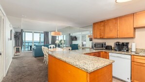 A modern kitchen with wooden cabinets, granite countertops, and a central island. There is a dining area with blue sofas and a large window overlooking a balcony. - Sunset Sips & Sandy Trips | Your Family’s Bayfront OCMD Escape (Ocean City)