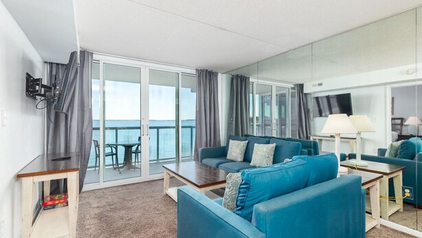 A modern hotel room with a balcony, a sofa set, a small table, and a television. - Sunset Sips & Sandy Trips | Your Family’s Bayfront OCMD Escape (Ocean City)