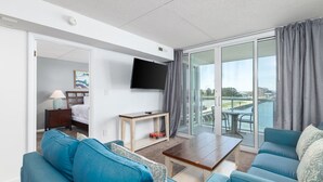 A modern hotel room with a living area featuring a sofa set, a flat-screen TV, and a balcony with a view of the water. - Sunset Sips & Sandy Trips | Your Family’s Bayfront OCMD Escape (Ocean City)