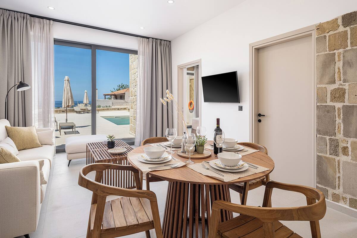 A dining area with a view of a pool and beach, wooden furniture, and a flat-screen TV on the wall.