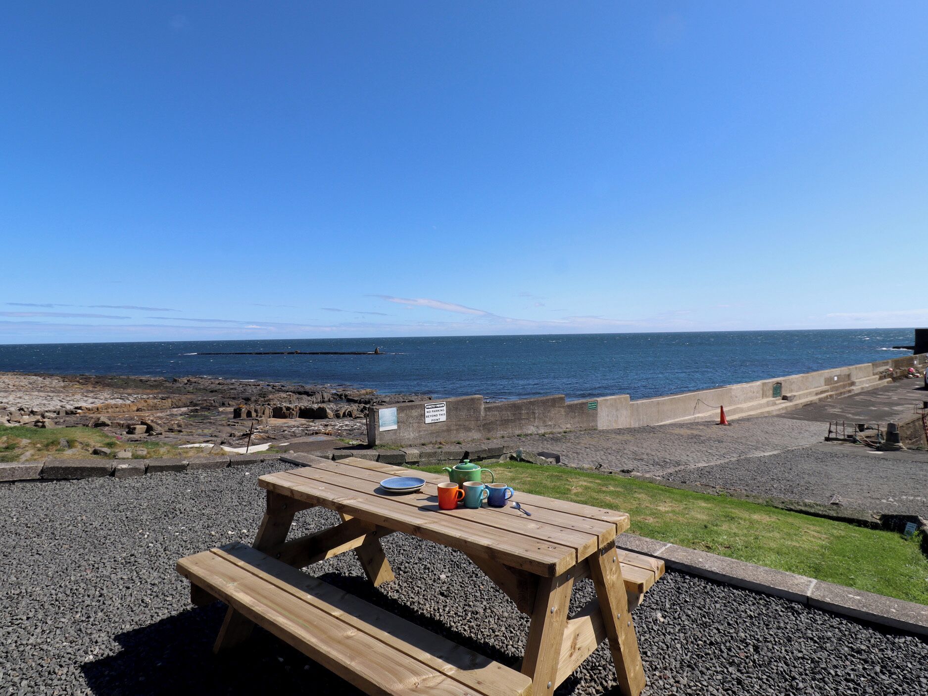 Ett picknickbord i trä med en tallrik, en mugg och en kopp på, placerat med utsikt över en stenstrand och med fri sikt ut över havet.