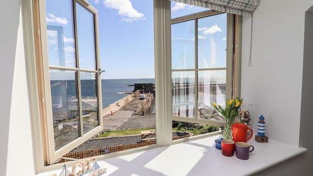 A room with a large window overlooking a coastal area, a white shelf with decorative items, and a sign reading "BEACH". - Puffin Cottage (Alnwick)