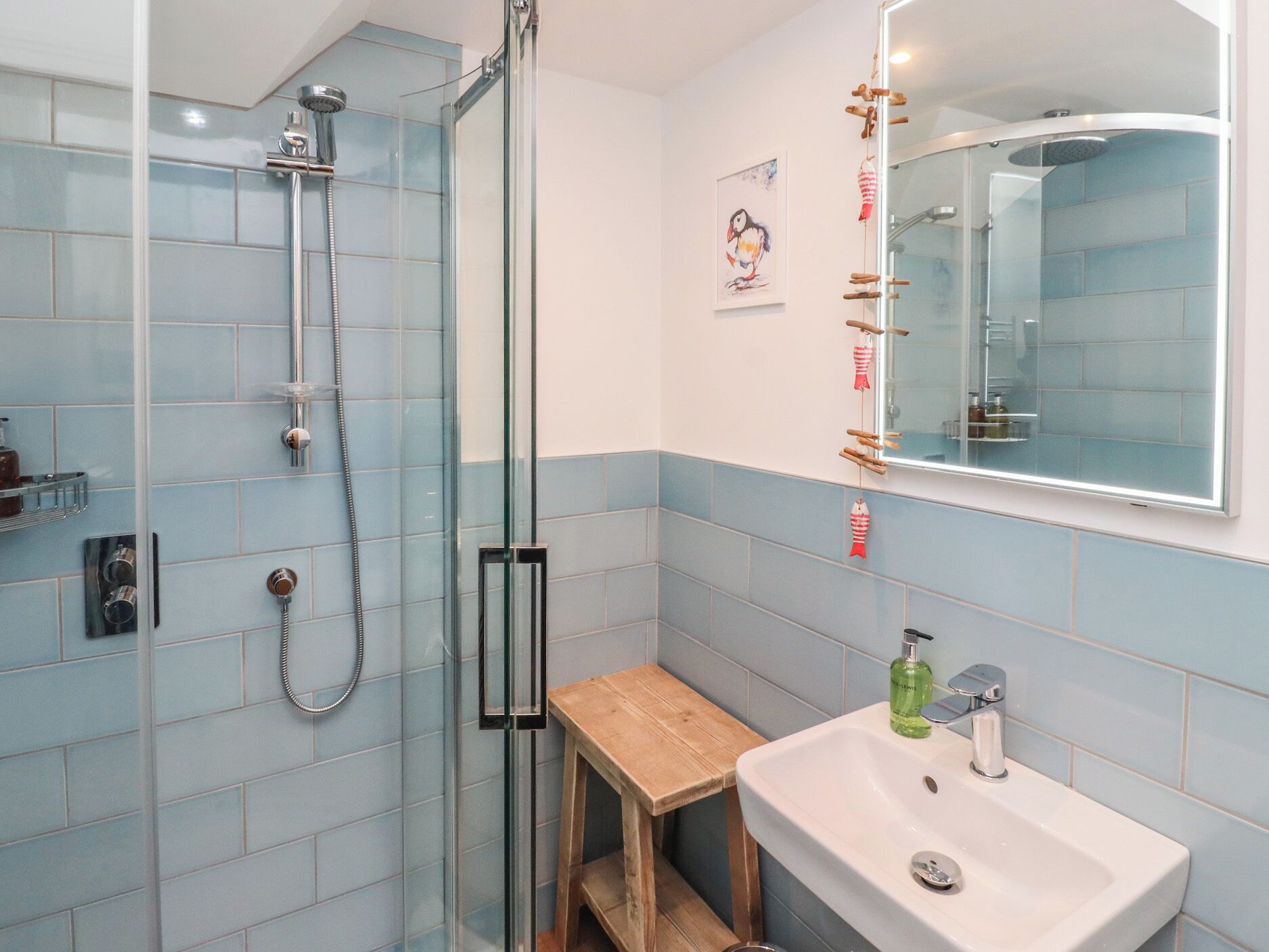 Een badkamer met een glazen douchecabine, een houten krukje, een wastafel met spiegel en een handdoekhouder aan de muur.