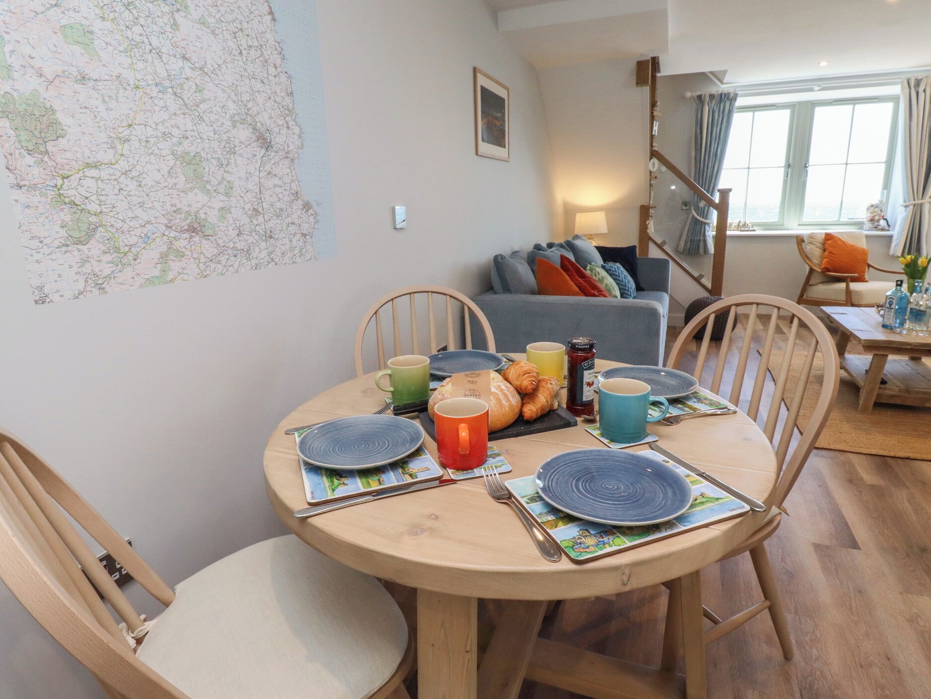 Een eethoek met een ronde houten tafel, gedekt voor vier personen, omringd door houten stoelen. Op de muur hangt een kaart, en op de achtergrond staat een bank.