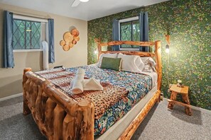 A room with a wooden bed, a window with curtains, a wall with a floral pattern, and a small wooden stool.