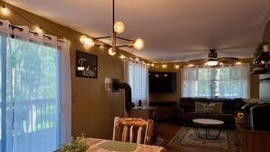 A cozy living room with a sofa, a coffee table, and a TV mounted on the wall. There is a dining area with a table and chairs, and a kitchen area with a counter and cabinets. - Throwback Lodge - Hot Tub - Pet Friendly (Gouldsboro)