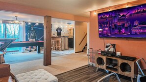 A modern living room with a pool table, bar area, and a large TV screen displaying a cityscape. - Throwback Lodge - Hot Tub - Pet Friendly (Gouldsboro)