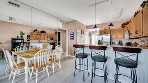 A spacious kitchen with a dining area, featuring a wooden table and chairs, a bar area with stools, and wooden cabinets. - New For 2025! Two Bedroom Bayfront Condo On 79th St (Ocean City)