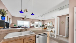A modern kitchen with a built-in dishwasher, a large sink, and wooden cabinets. There is a dining area with a white table and chairs, and a ceiling fan above. - New For 2025! Two Bedroom Bayfront Condo On 79th St (Ocean City)