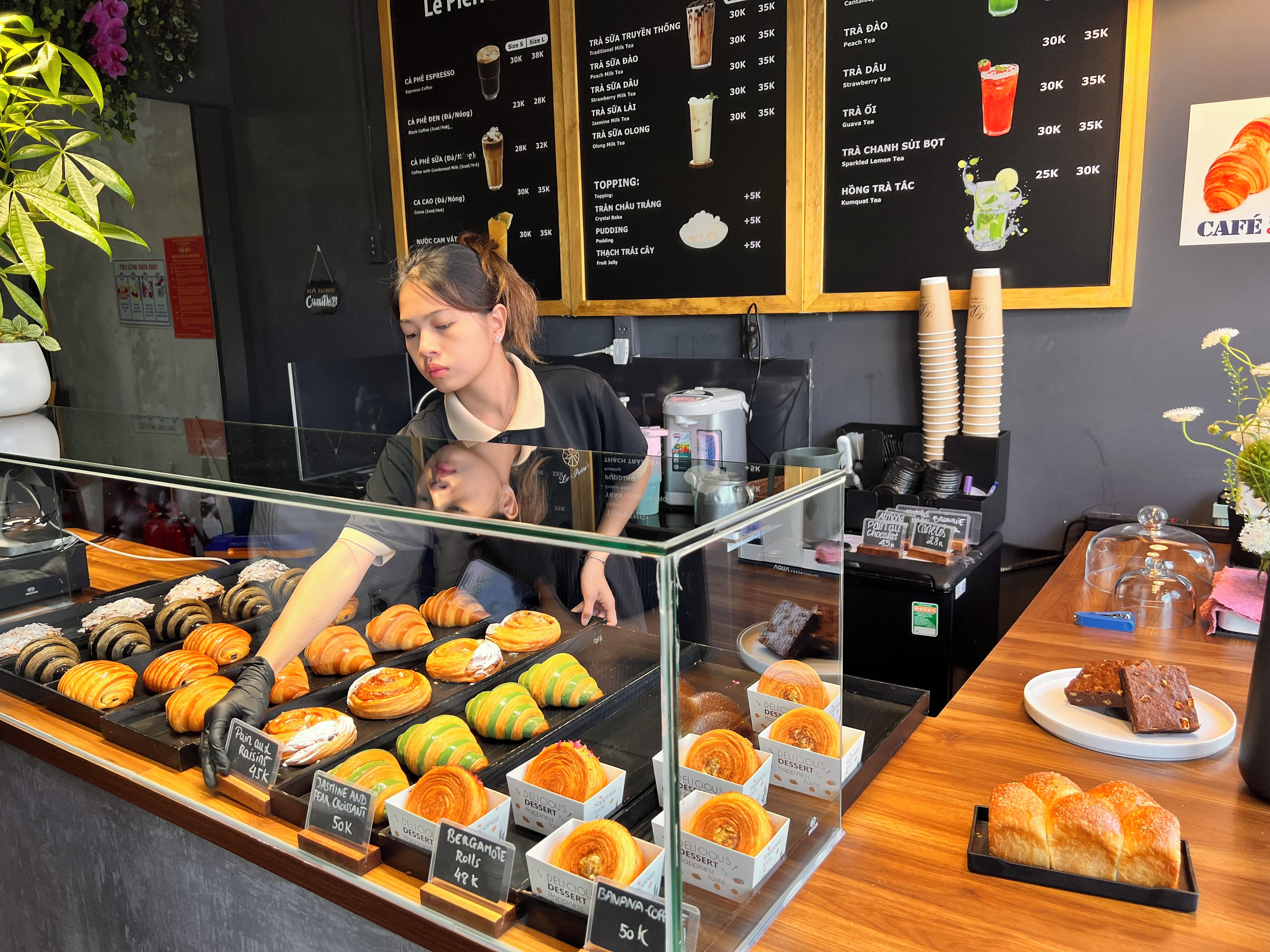 A bakery counter with various pastries and desserts displayed in clear containers, a menu board above, and a coffee machine to the side.