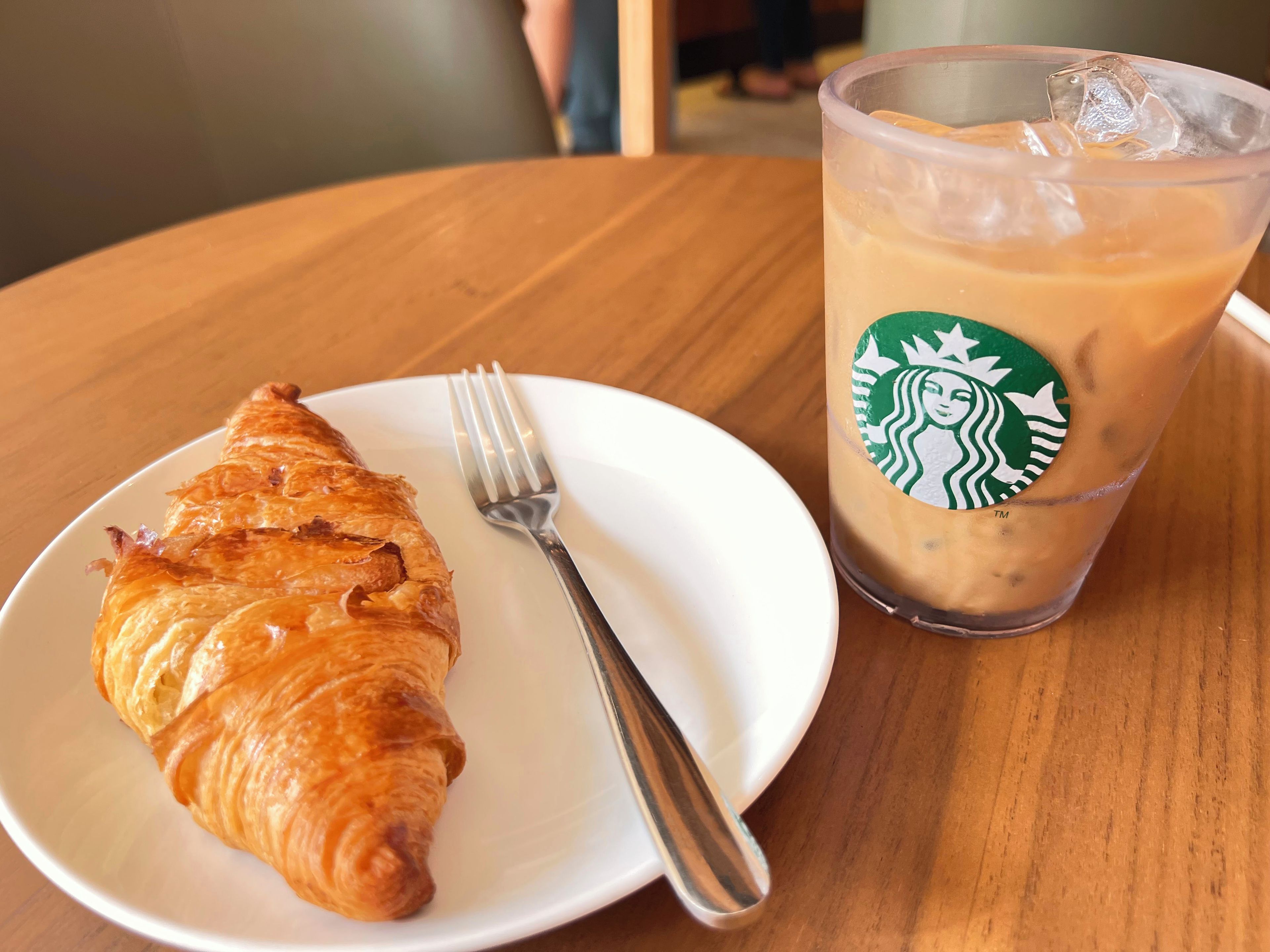 A croissant on a white plate with a fork beside it, and a Starbucks iced coffee cup with a straw.