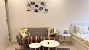 Ein modernes Wohnzimmer mit einem grauen Sofa, gemusterten Kissen, zwei kleinen weißen Tischen, einem großen Teppich mit Muster und einem Bett mit Blumenmuster auf der Bettwäsche.
