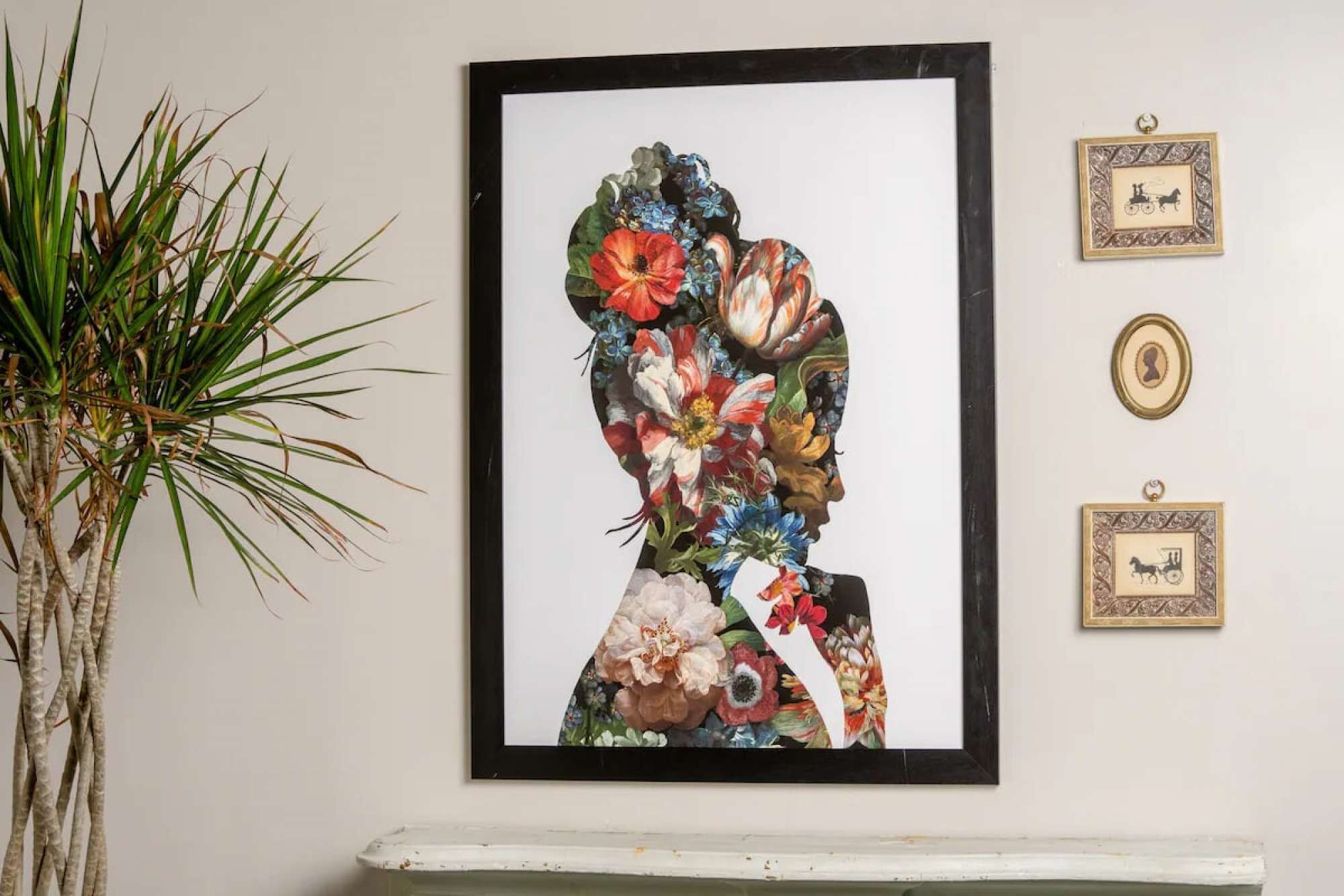 A framed floral artwork on a wall, a potted plant to the left, and two small framed pictures above a white shelf.