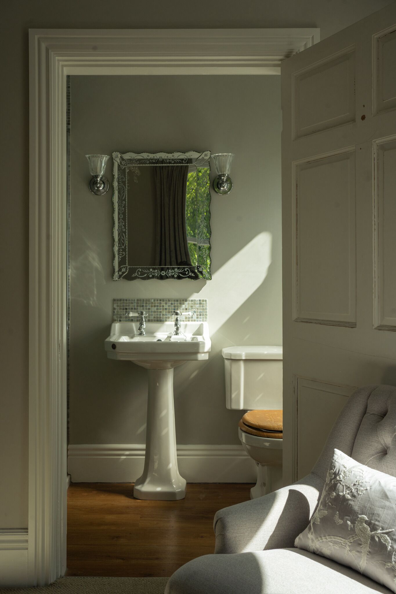 A bathroom with a pedestal sink, a toilet, and a chair with a cushion.