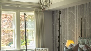 A bedroom with a four-poster bed, a chandelier, a window with curtains, a radiator, and a bedside table with a lamp. - Outwood House (Leeds)
