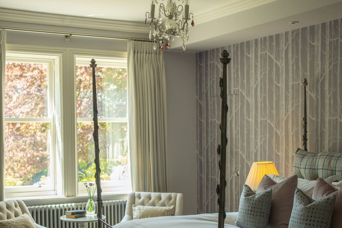 Ein Schlafzimmer mit Himmelbett, einer Chandelierleuchtre, einem Fenster mit Vorhängen, einem Heizkörper und einem Nachttisch mit Lampe.