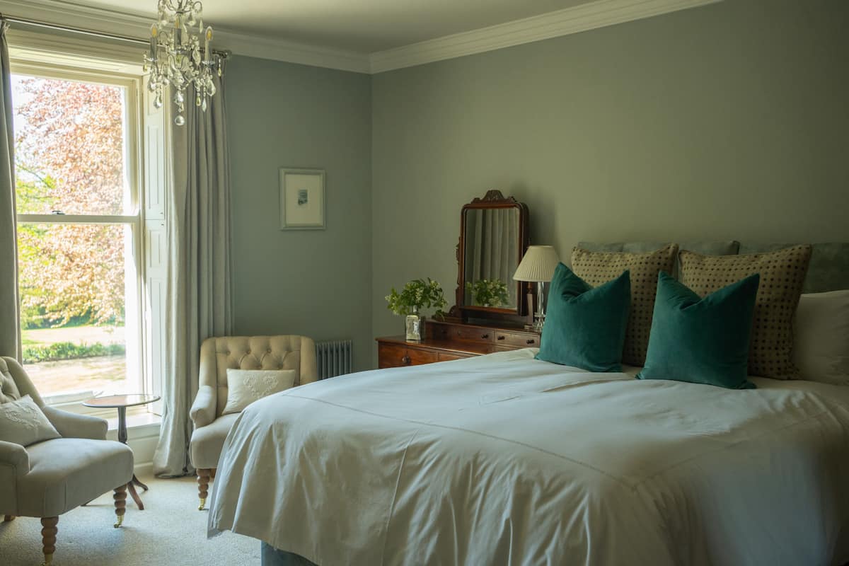 Ein Schlafzimmer mit einem großen Bett, einem Sessel, einem Schminktisch und einem Fenster mit Vorhängen.