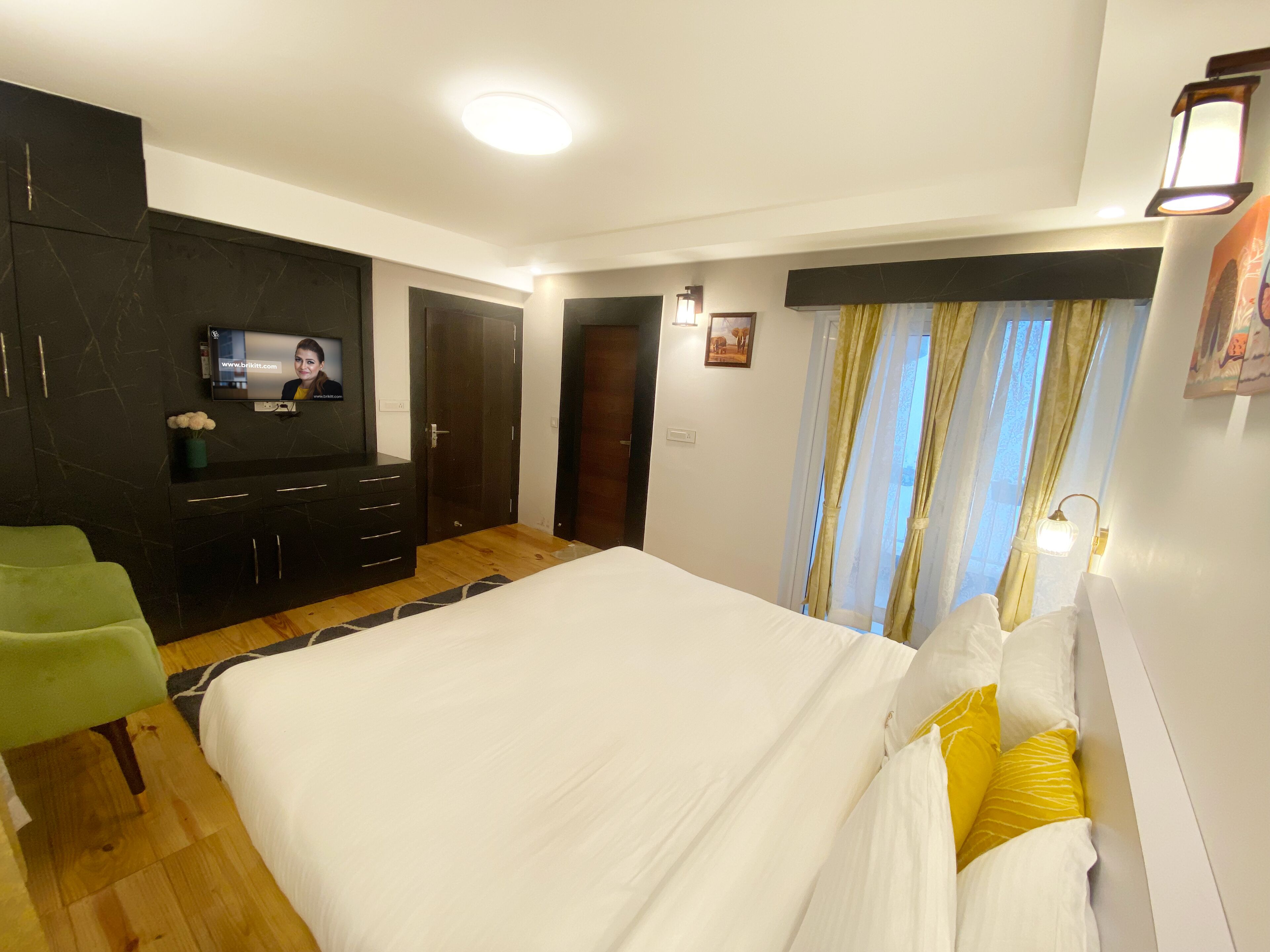 A bedroom with a large bed, a TV mounted on the wall, a green chair, and a wooden floor.