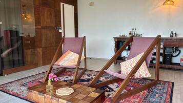 A room with two wooden chairs with cushions, a coffee table, a patterned rug, and a kitchen area with a water dispenser.