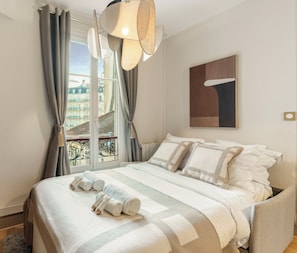 Habitación de hotel con cama, vista de edificios a través de una ventana, lámpara colgante y un cuadro en la pared.