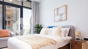 A modern bedroom with a large bed, a nightstand, a sofa, and a view of the city through large windows.