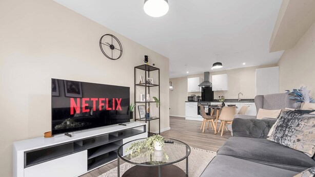 Uma sala de estar moderna com uma televisão de tela plana, uma mesa de centro, um sofá cinza e uma cozinha ao fundo.