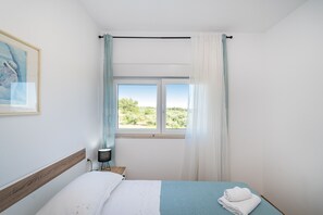 Ein Schlafzimmer mit Bett, Vorhängen am Fenster, einer Nachttischlampe und einem gerahmten Bild an der Wand.