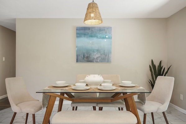 Un coin repas comprenant une table en verre, deux chaises, un luminaire suspendu en fibres tressées et un grand tableau abstrait accroché au mur.