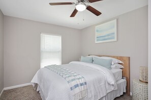 A bedroom with a bed, a ceiling fan, a window with blinds, and a wall-mounted picture.