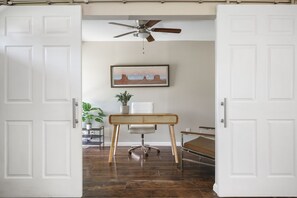An open door leading to a room with a ceiling fan, a desk with a chair, a bed, and a framed picture on the wall.