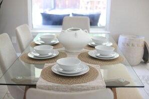 Une table à manger sur laquelle sont disposés un pot en céramique blanche, quatre tasses et sous-tasses blanches, le tout posé sur une table en verre agrémentée de sets de table en fibres tressées.