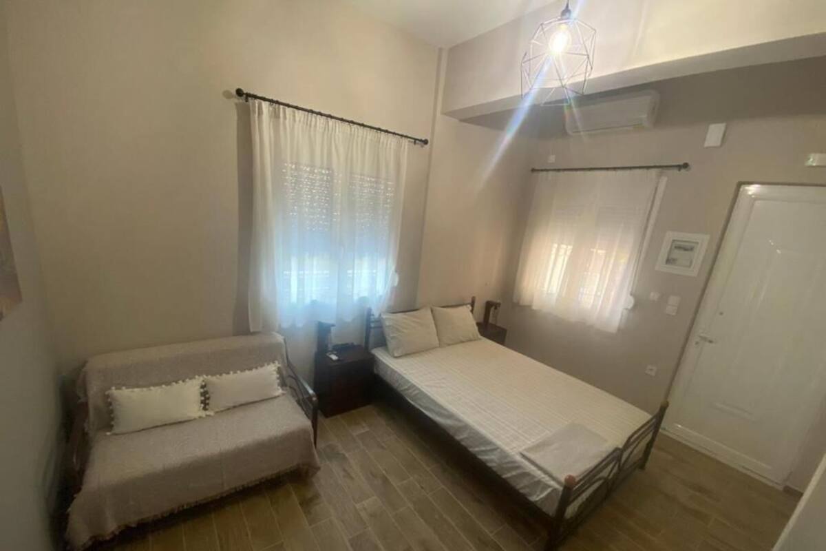 A small, well-lit bedroom with a bed, a nightstand, a chair, and a window with sheer curtains.