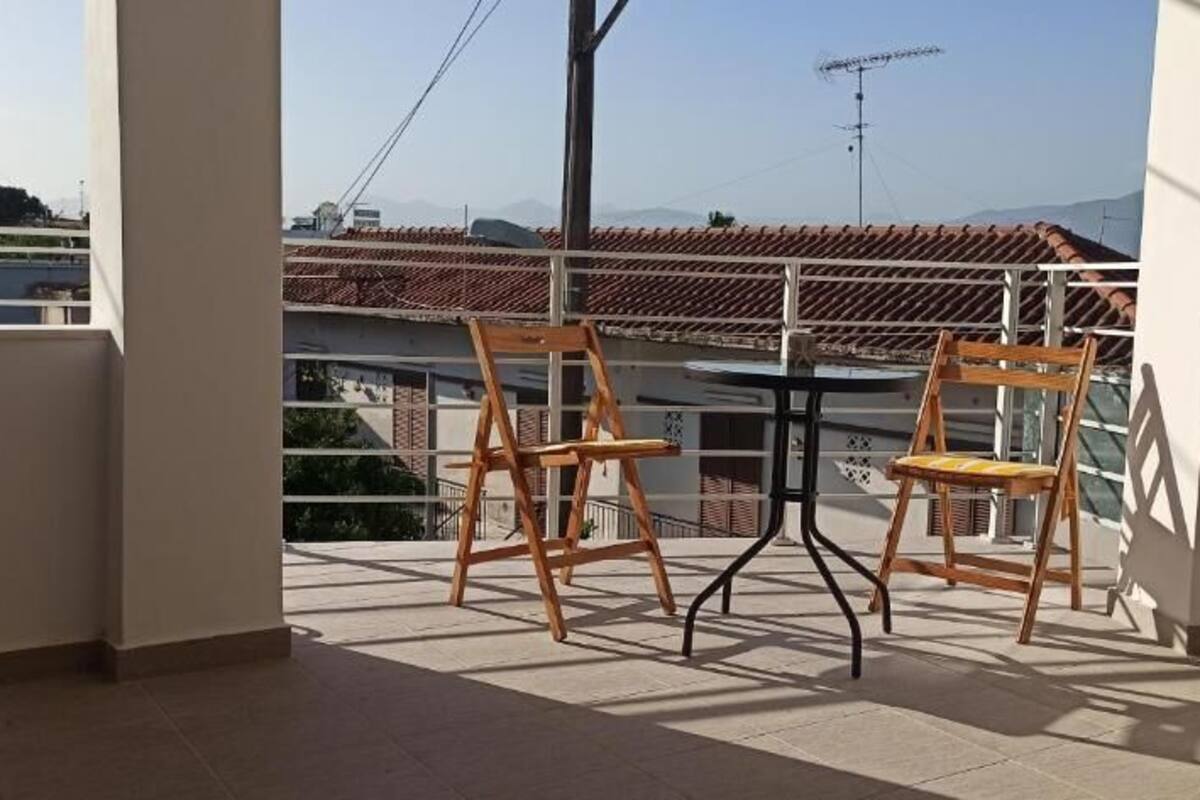 A balcony with two wooden chairs and a table, overlooking a tiled roof and buildings.