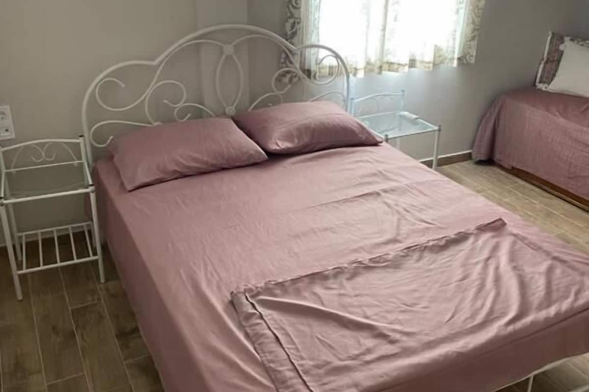 A neatly made bed with a pink bedspread and pillows, a white metal headboard, a bedside table with a lamp, and a window with curtains.