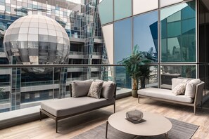 Un salón moderno con un sofá y un sillón, una mesa redonda y un amplio ventanal que ofrece vistas a un paisaje urbano y una estructura esférica singular.