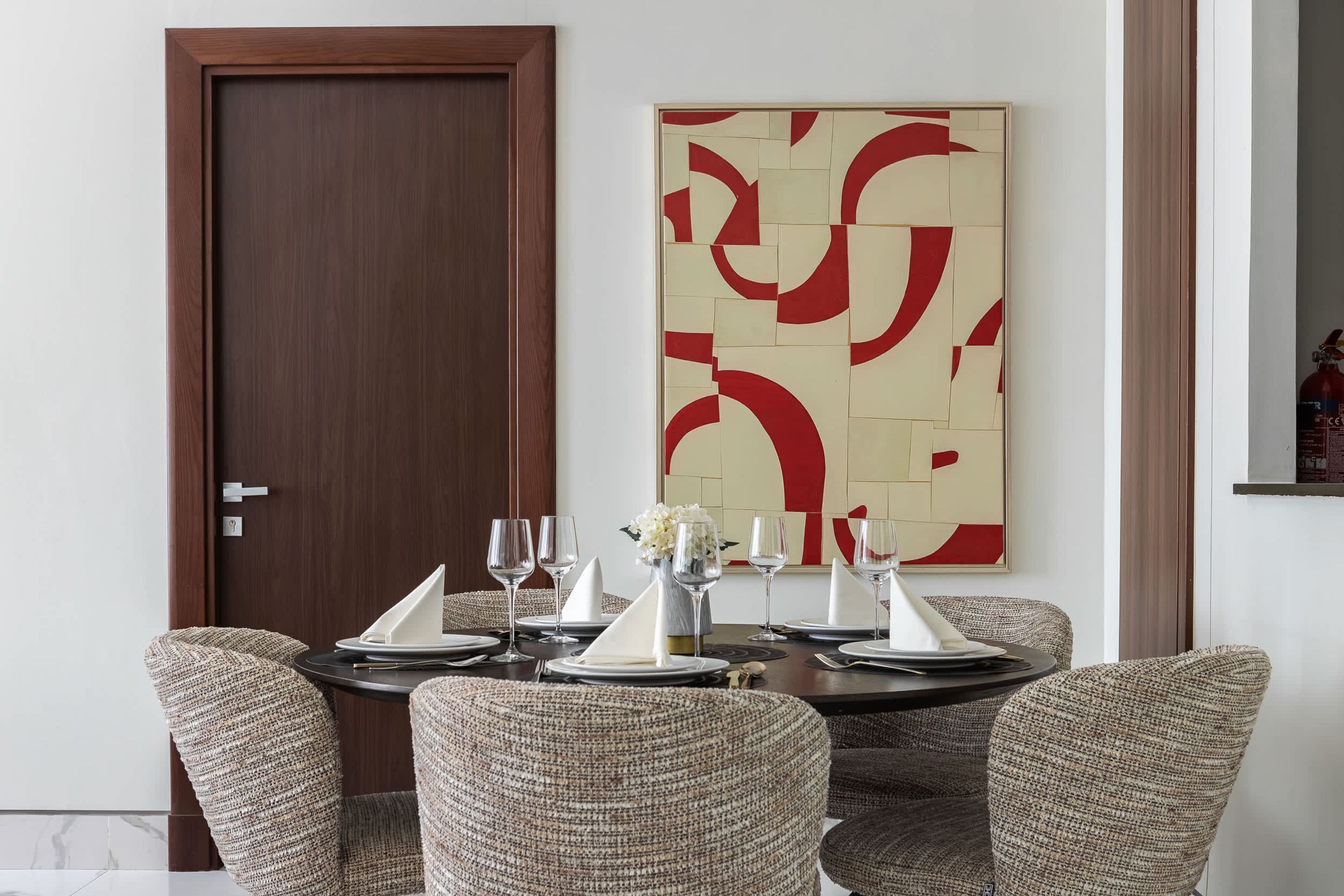 A dining area with a round table set for four, surrounded by upholstered chairs. A large abstract painting is on the wall above the table.